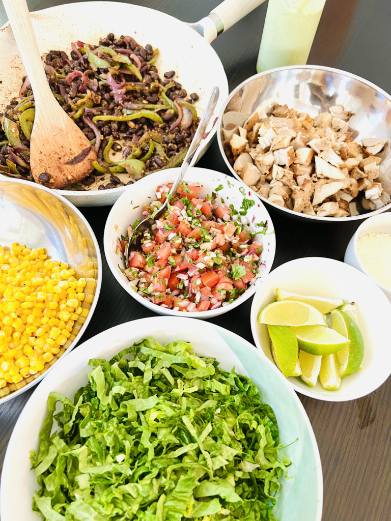 Bowls of lettuce, pico de gallo, corn, beans and peppers, meat, lime
