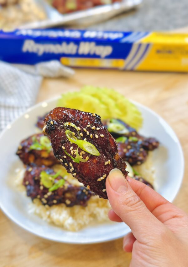 Single honey soy chicken wing held by hand in focus with plate of wings in background