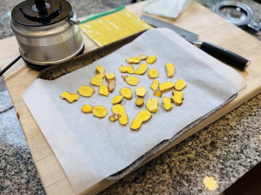 Sliced Ginger in a parchment paper in a flat pan and knife on the side, food processor on the left top and a Ginger in a zip lock