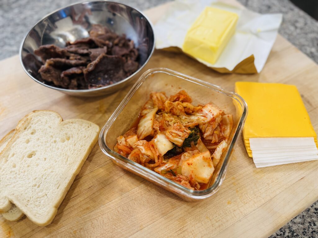Ingredients for Kimchi Grilled Cheese with Bulgogi — sourdough bread, kimchi, cooked bulgogi, American cheese slices, butter, and sesame oil arranged on a surface