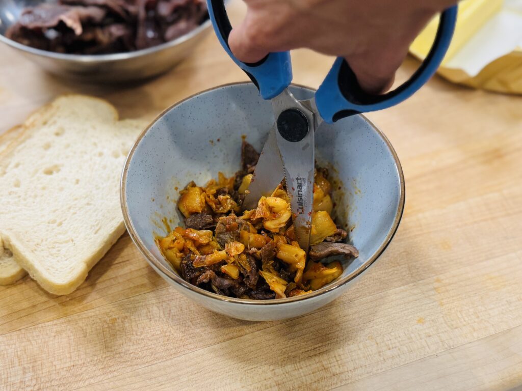 Kitchen scissors chopping caramelized kimchi and bulgogi together in a bowl into bite-sized pieces