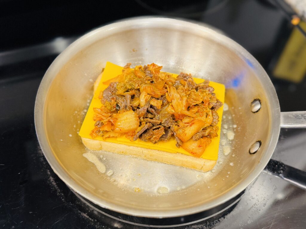 Kimchi grilled cheese being assembled in a skillet — sourdough bread with American cheese, kimchi, and bulgogi filling layered inside
