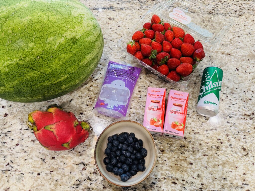 Hwachae ingredients flat lay — fresh watermelon, strawberries, dragonfruit, blueberries, coconut jelly, strawberry milk boxes, and Chilsung Cider cans arranged on a surface