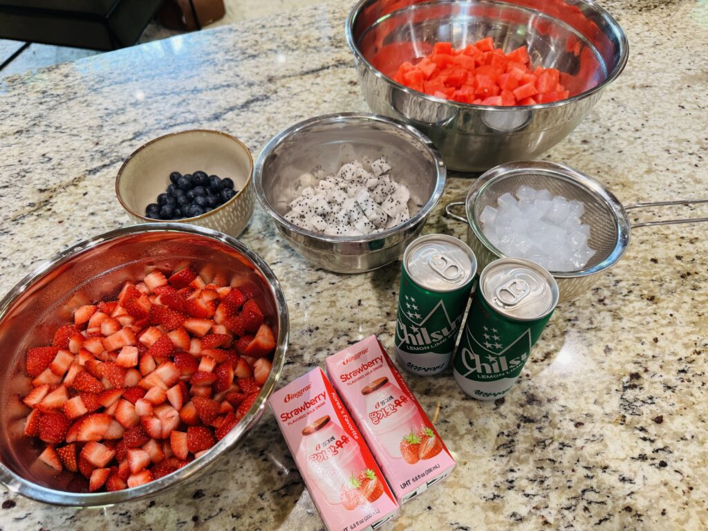 Hwachae ingredients flat lay with fruit chopped and prepped — fresh watermelon, strawberries, dragonfruit, blueberries, coconut jelly, strawberry milk boxes, and Chilsung Cider cans arranged on a surface