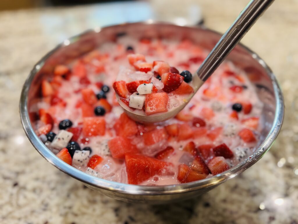 Ladle lifting a scoop of hwachae Korean fruit punch from a large bowl, showing fresh watermelon, strawberries, dragonfruit, blueberries, and coconut jelly