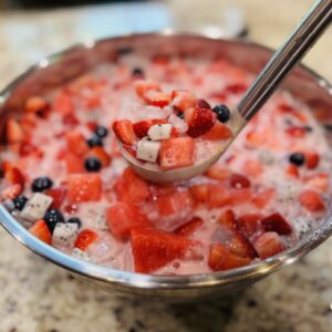 Ladle lifting a scoop of hwachae Korean fruit punch from a large bowl, showing fresh watermelon, strawberries, dragonfruit, blueberries, and coconut jelly