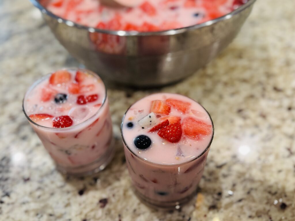 Individual glass serving of hwachae Korean fruit punch with fresh fruit and coconut jelly, with the full hwachae bowl visible in the background