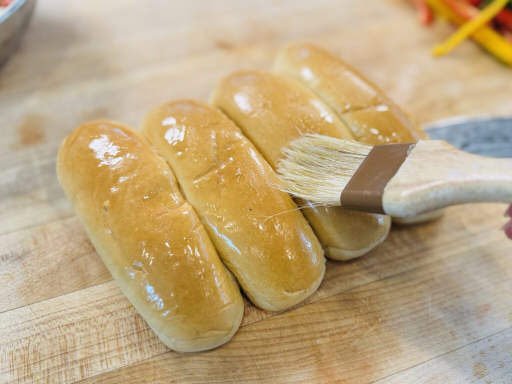 Melted butter being brushed onto the outside of hot dog buns with a pastry brush before toasting in a pan