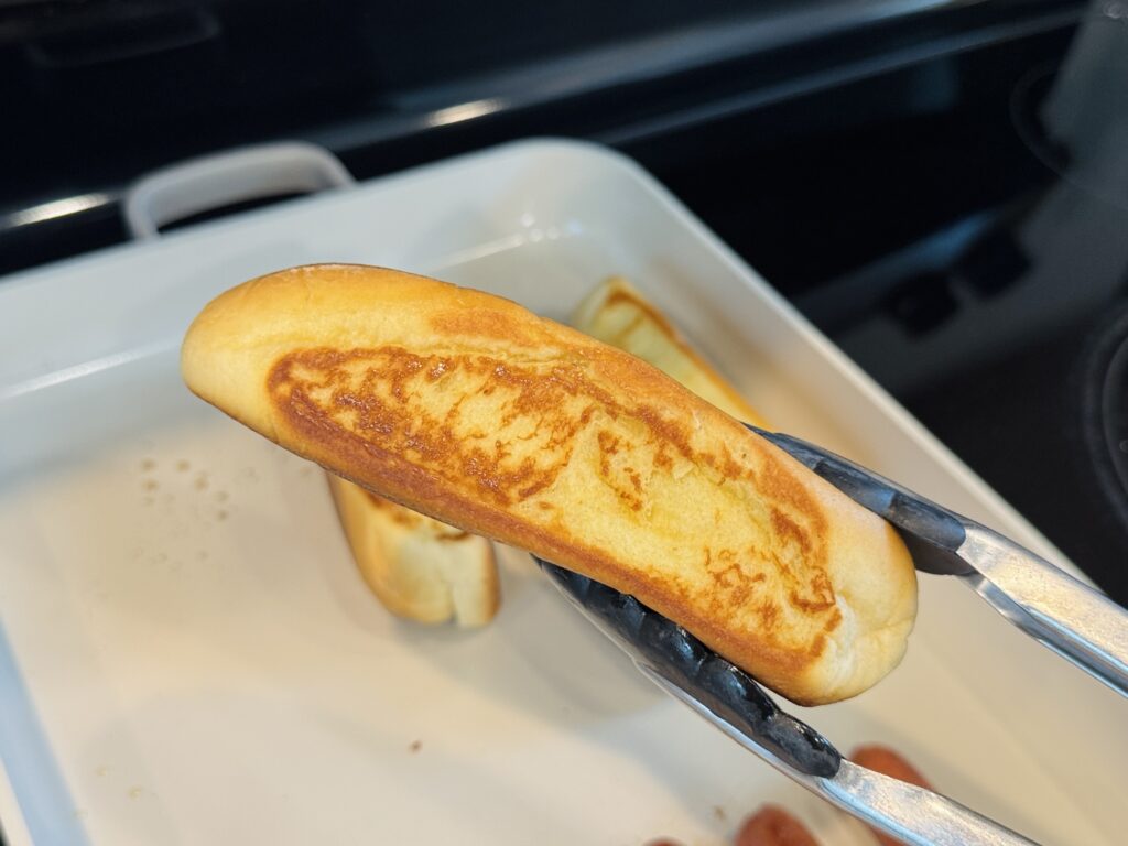 Melted butter being brushed onto the outside of hot dog buns with a pastry brush before toasting in a pan