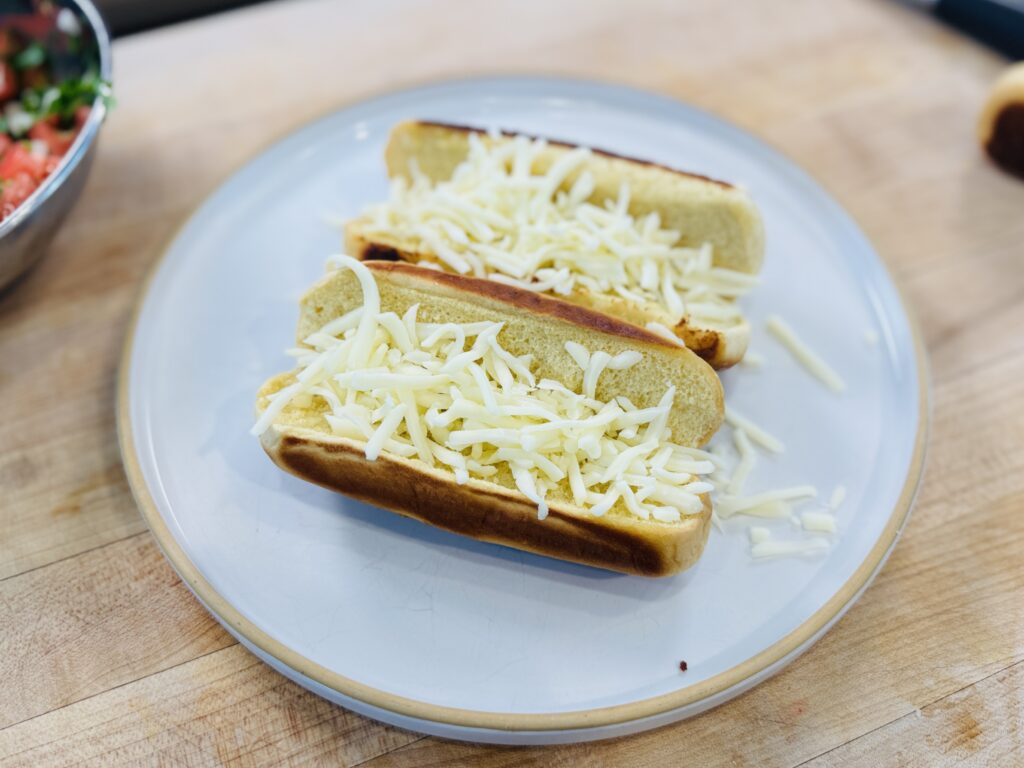 Shredded mozzarella cheese sprinkled inside toasted hot dog buns before the hot dog is added, ready to be melted