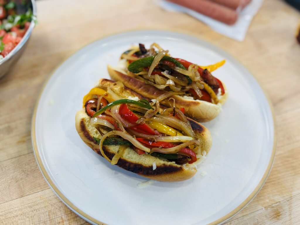 Mexican hot dogs being assembled — caramelized bell peppers and onions being layered over a hot dog in a toasted bun