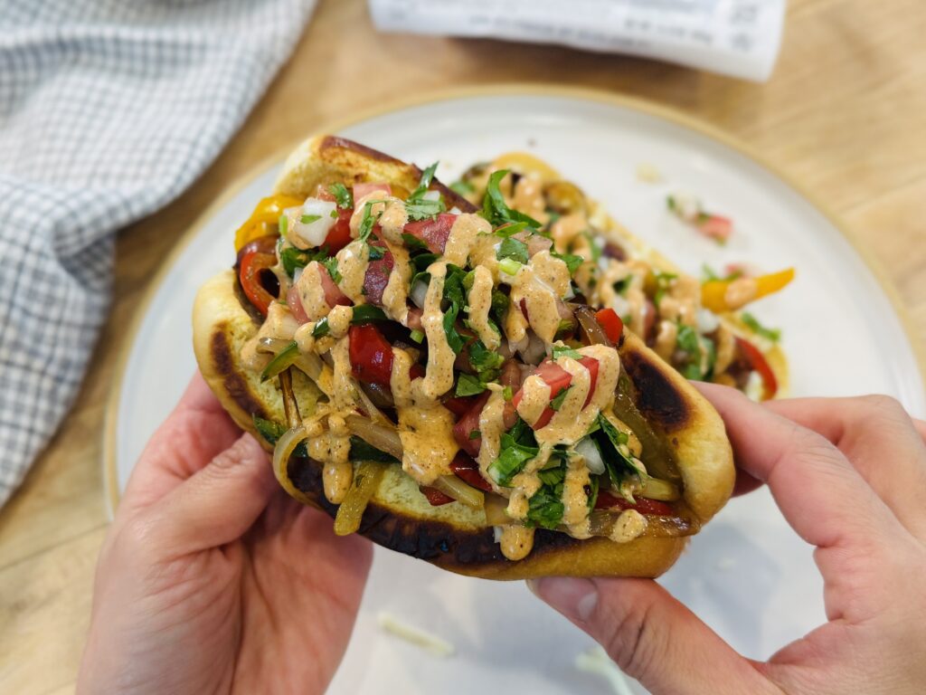Mexican hot dogs fully assembled on toasted buttered buns with melted cheese, caramelized peppers and onions, fresh pico de gallo, and homemade creamy sauce drizzled over the top