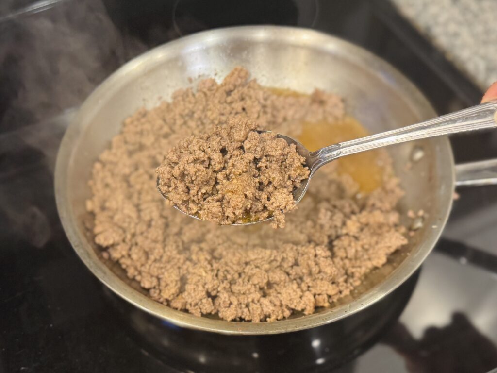 Close-up of seasoned ground beef cooking in a skillet, broken into fine crumbles, lifted with a large spoon to show texture
