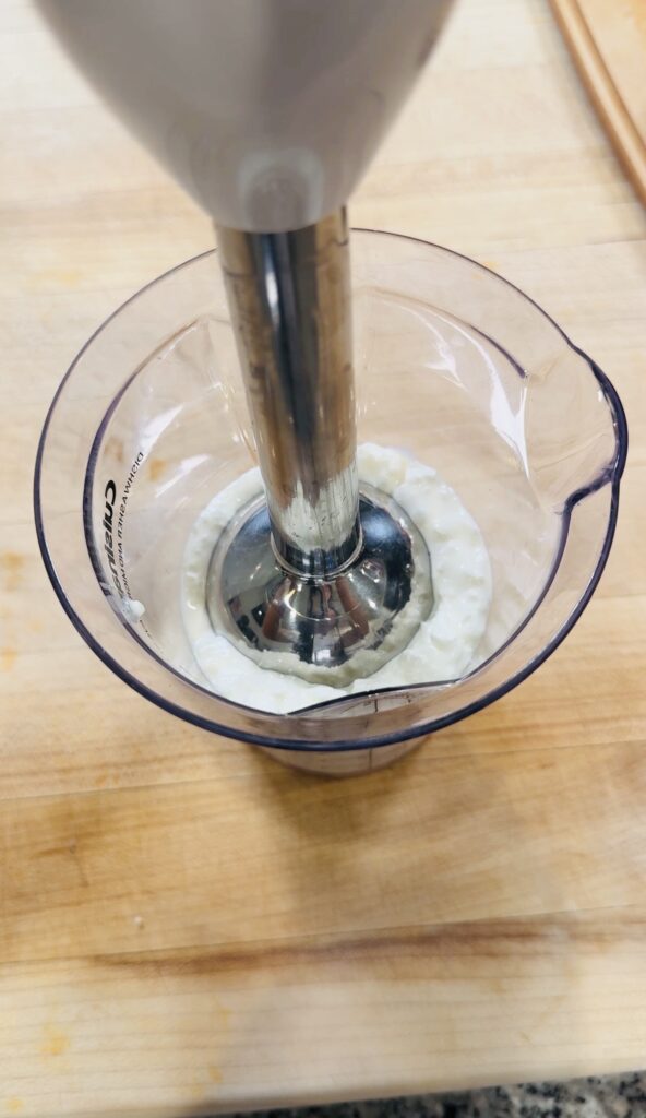 Cottage cheese and honey being blended with an immersion blender in a cup until smooth and whipped in texture.