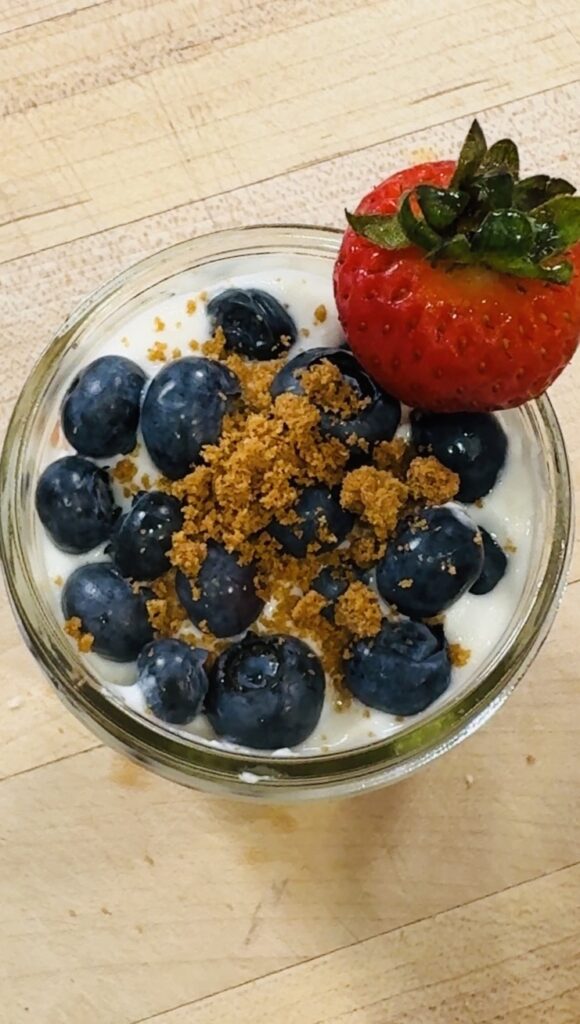 Overhead shot of high protein berry trifle in a mason jar — fresh blueberries as the top layer, garnished with crushed Biscoff cookies and a strawberry on the rim