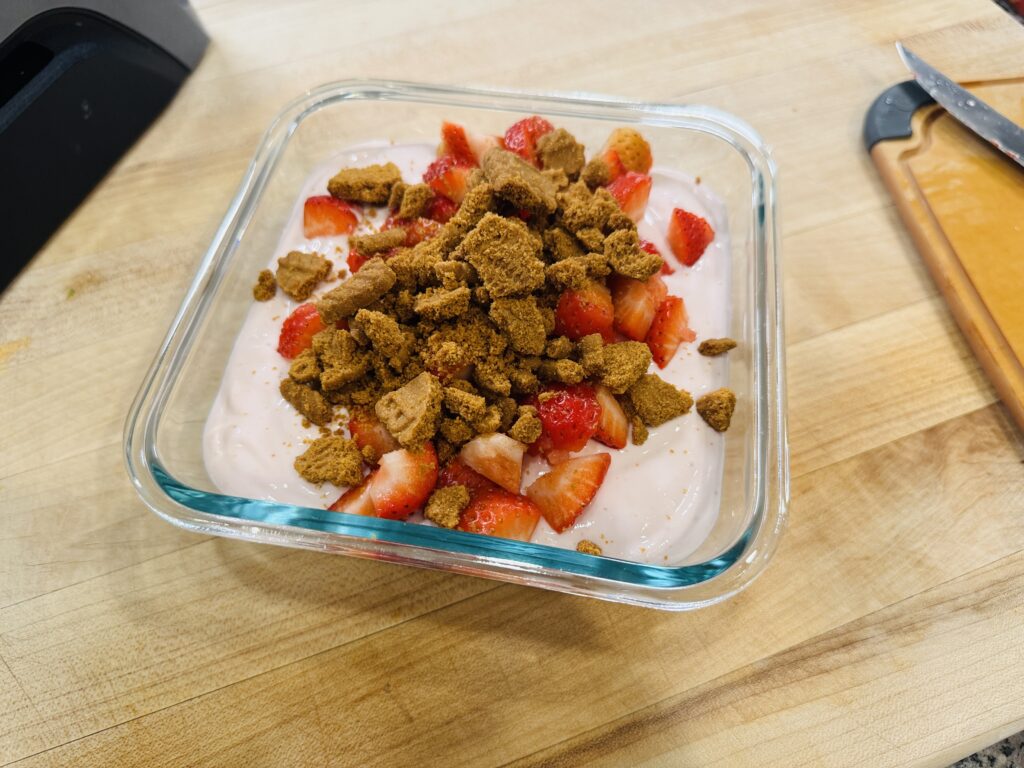 Blended strawberry cottage cheese ice cream mixture in an airtight glass container, topped with crushed Biscoff cookies and fresh strawberries, ready to go into the freezer