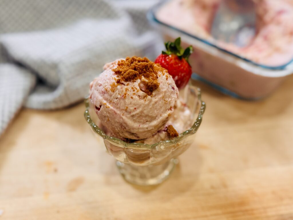 Strawberry Biscoff cottage cheese ice cream scooped into a bowl, garnished with crushed Biscoff cookies and a fresh strawberry on the rim