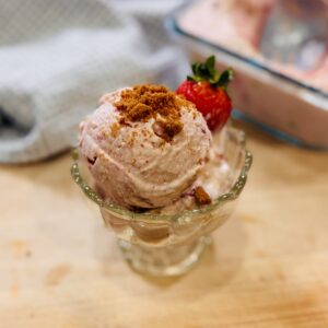 Strawberry Biscoff cottage cheese ice cream scooped into a bowl, garnished with crushed Biscoff cookies and a fresh strawberry on the rim