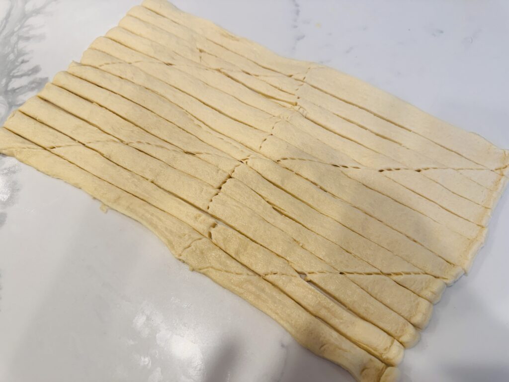 Crescent dough rolled out and cut lengthwise into long half-inch strips with a pizza cutter, ready to wrap around hot dogs.