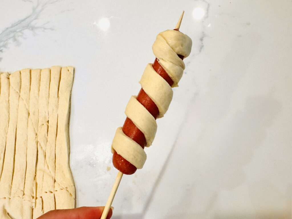 Close-up of a single hot dog on a wooden skewer wrapped in crescent dough strips in a spiral pattern, ready to go into the oven