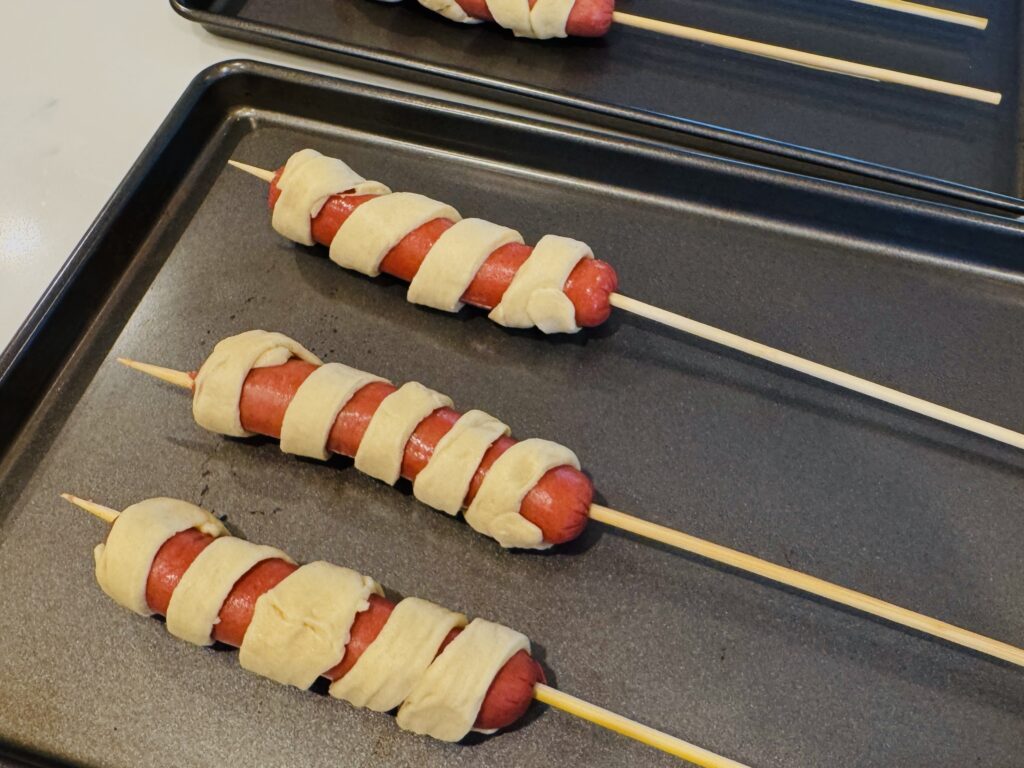 Eight crescent dough-wrapped hot dogs on wooden skewers laid out on a parchment-lined baking sheet, ready to go into the oven.