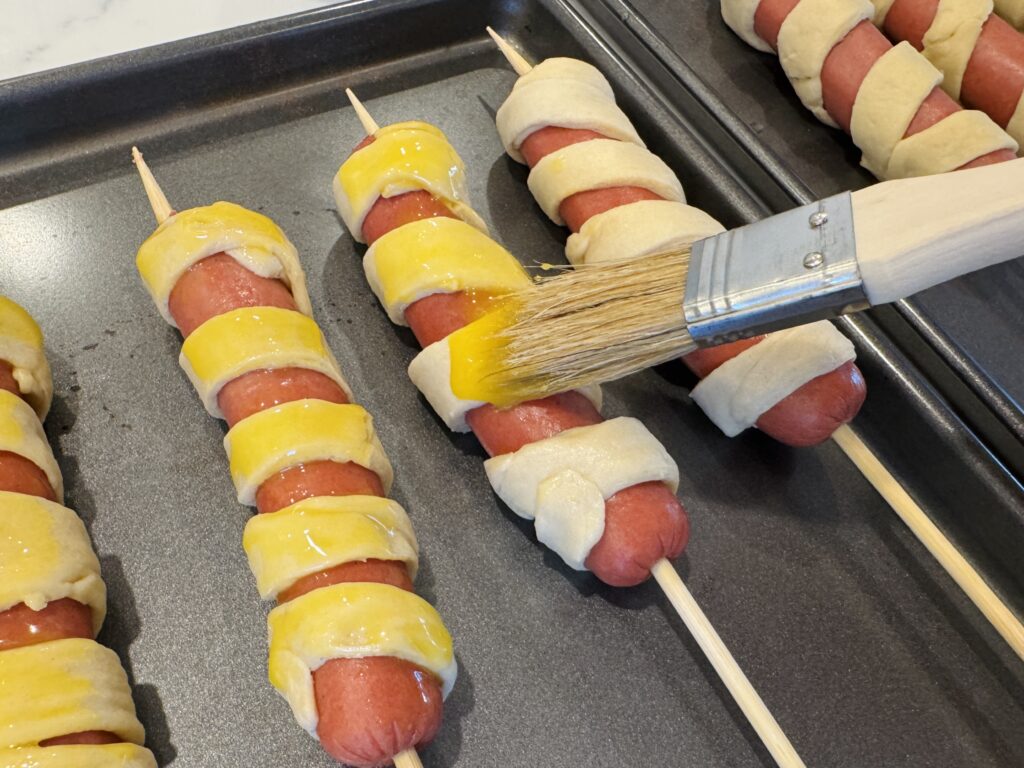 Egg wash being brushed onto crescent dough-wrapped firecracker hot dogs on a baking sheet before going into the oven