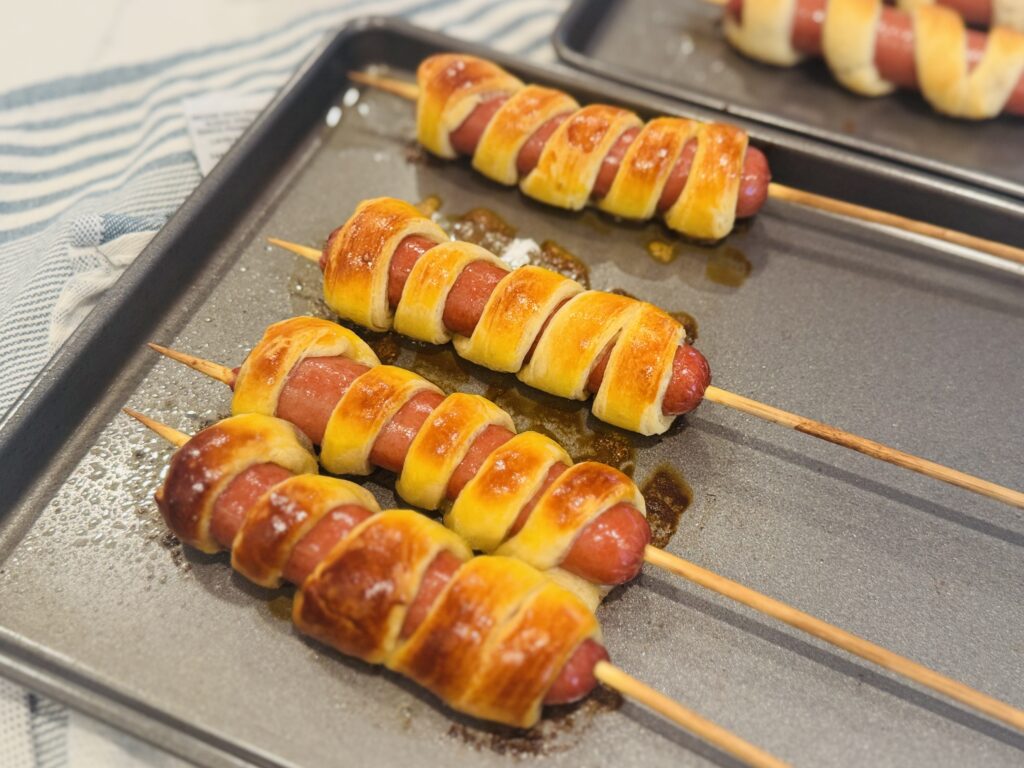 Crescent dough-wrapped firecracker hot dogs on wooden skewers just out of the oven, golden brown on a parchment-lined baking pan