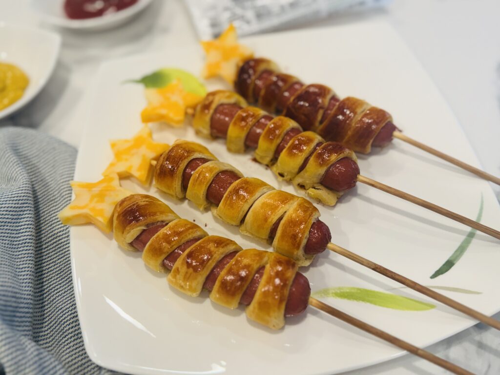 Four firecracker hot dogs laid out on a plate — crescent dough-wrapped hot dogs on wooden skewers topped with cheddar cheese stars