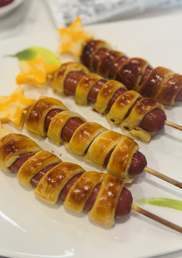 Four firecracker hot dogs laid out on a plate — crescent dough-wrapped hot dogs on wooden skewers topped with cheddar cheese stars