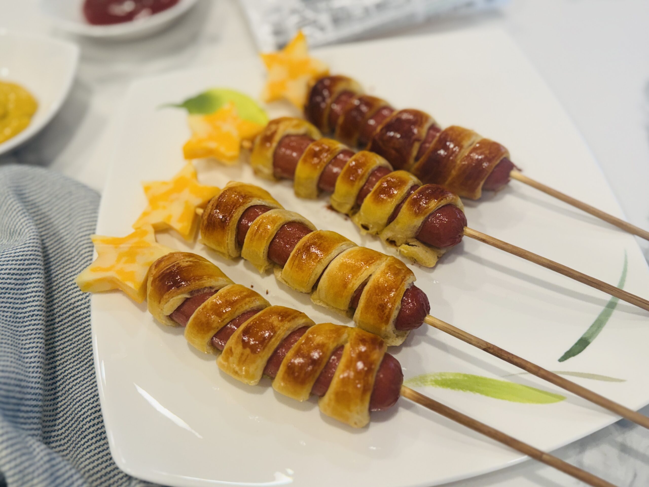 Four firecracker hot dogs laid out on a plate — crescent dough-wrapped hot dogs on wooden skewers topped with cheddar cheese stars