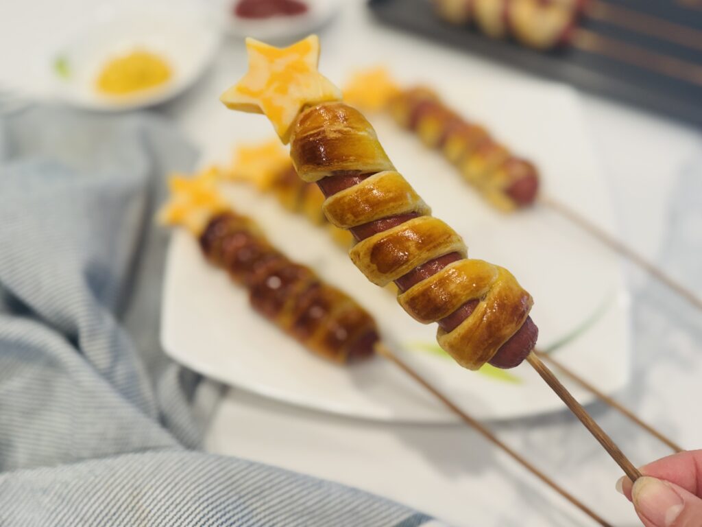 Close-up of one firecracker hot dog on a wooden skewer with a cheddar cheese star on top, remaining hot dogs plated in the background