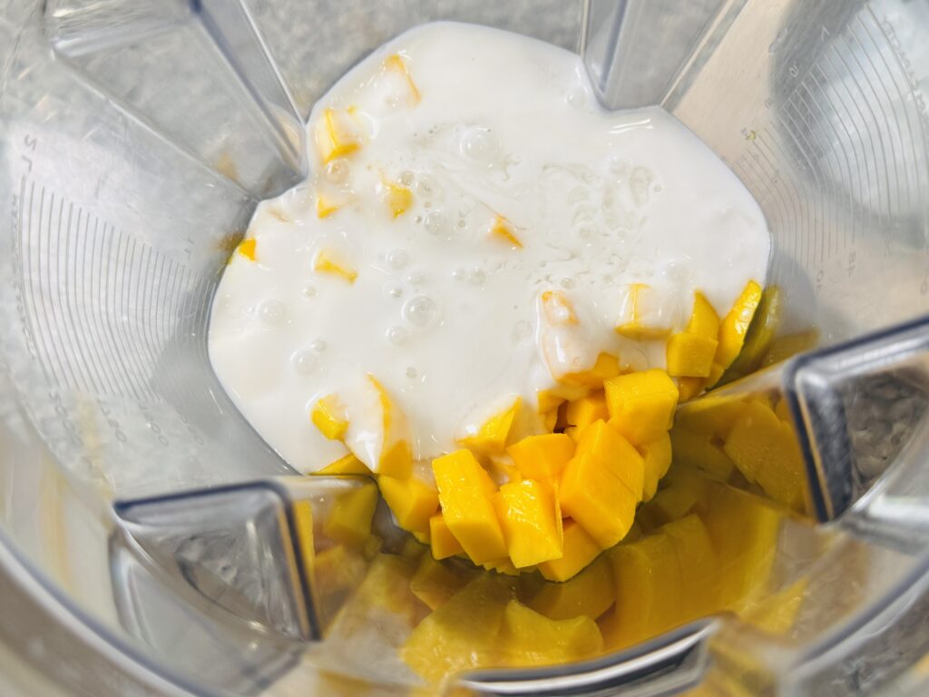 Chopped mangoes and coconut milk being poured into a blender with chopped mango