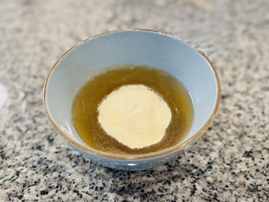 Beef gelatin powder being scooped and added on top of a bowl of apple juice, not yet whisked in