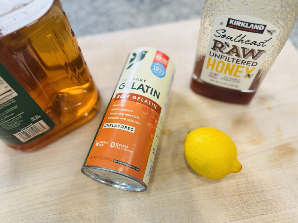 Overhead flat lay of the four ingredients for apple protein gummies: apple juice, gelatin, honey, and lemon