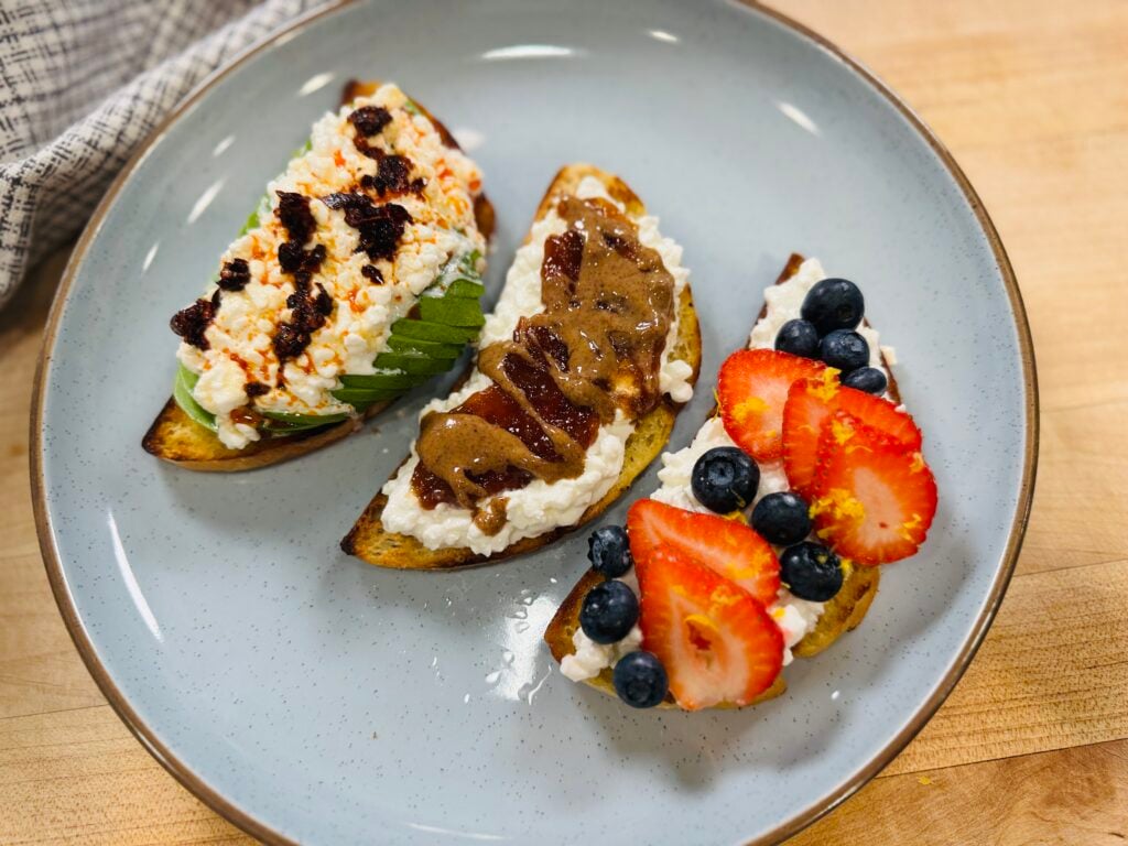 Three cottage cheese toasts on one plate: chili oil and honey with avocado, PB&J, and berries with lemon zest