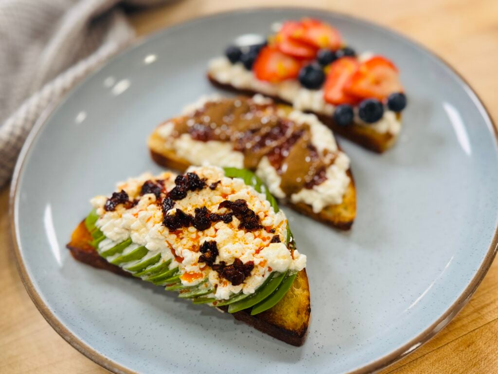 Close-up of three cottage cheese toast varieties on a plate, with the chili oil avocado toast in focus