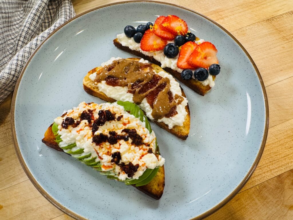 Overhead view of three cottage cheese toasts topped with avocado and chili oil, jam and peanut butter, and fresh berries